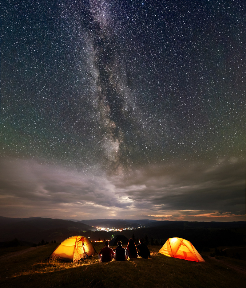 四名徒步旅行者坐在两个橙色帐篷营地, 晚上享受美丽的星空和银河。在远处的城市的灯光坐落在山之间图片