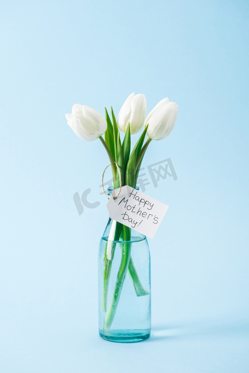 花束白色郁金香在玻璃花瓶与快乐的母亲天问候标签在蓝色背景图片