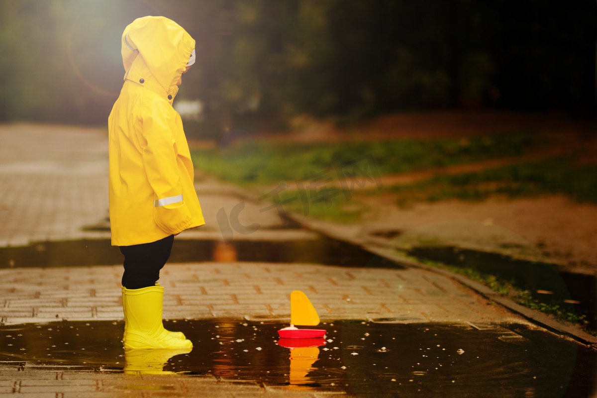 穿着黄色雨衣的男孩看着一艘漂浮在水坑中的玩具船。小婴儿在公园里散步.图片