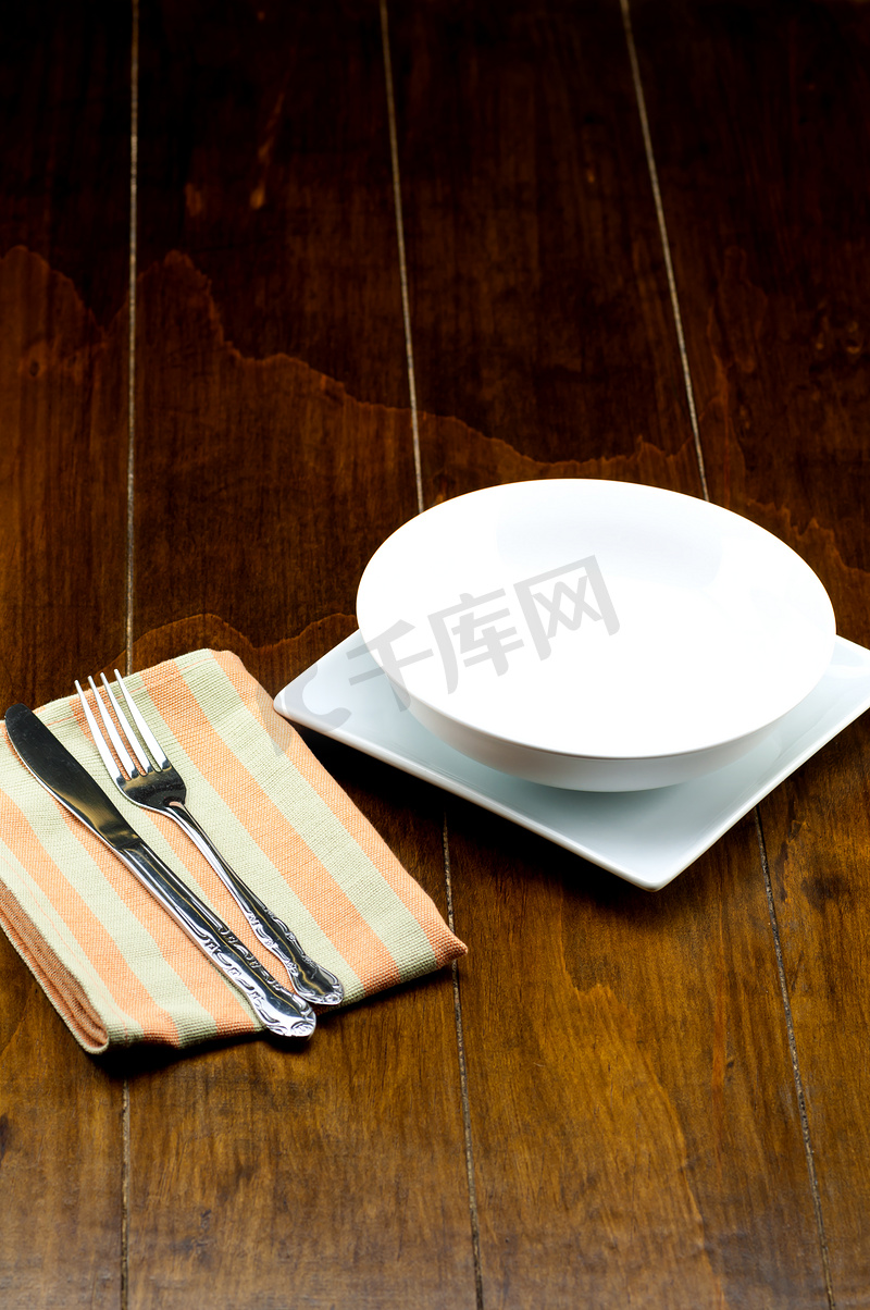 方形盘子上的空碗，餐巾上有叉子和刀子，在木质背景上。图片