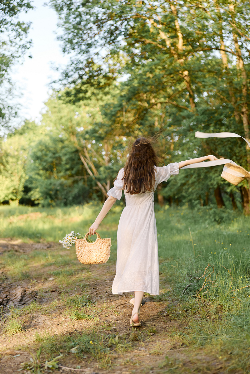 一位穿着夏长裙的女人沿着林间小路行走，手里拿着一顶柳条帽，上面有飘逸的丝带图片