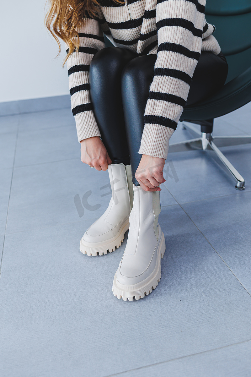 一位年轻漂亮的女人坐在椅子上，脚上穿着春季新款鞋款的白色皮靴。图片