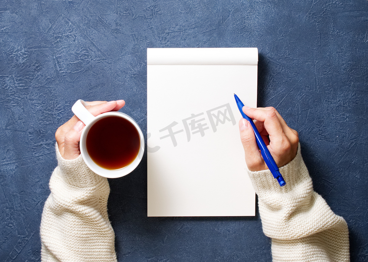 女人在深蓝色桌子上的笔记本上写字，手拿衬衫拿着铅笔，一杯茶，素描本，顶视图图片