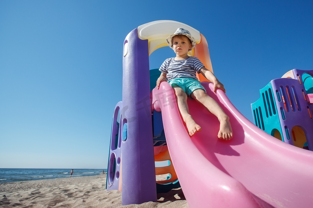海滩背景上彩色塑料滑梯，可爱的小男孩准备滑下来。图片