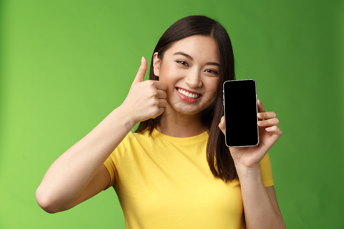 特写满意的可爱亚洲女孩给出积极的反馈，竖起大拇指显示智能手机屏幕，介绍很棒的新应用程序，推荐好的应用程序，吹嘘游戏中的击败分数图片