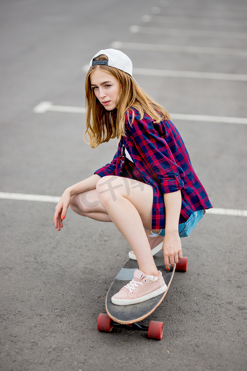 一个美丽的金发女孩穿着格子衬衫，帽子和牛仔短裤坐在停车场的长板上，一个深思熟虑的样子。运动和酷炫的风格.图片