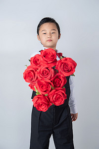 小孩自然摄影照片_情人节白天穿西装的帅气男孩室内手拿玫瑰花举向镜头摄影图配图