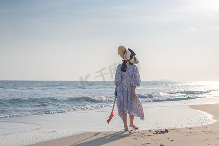 散步摄影照片_海边带帽子的女人下午穿碎花衣服的女人沙滩散步摄影图配图