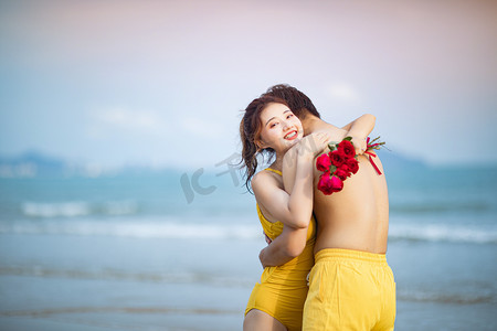 海边情侣摄影照片_爱情白天一对情侣沙滩拿着玫瑰花拥抱摄影图配图