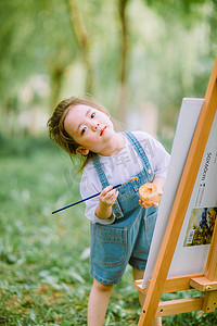 摄影照片_艺术启蒙白天画画的小女孩户外草坪歪头摄影图配图