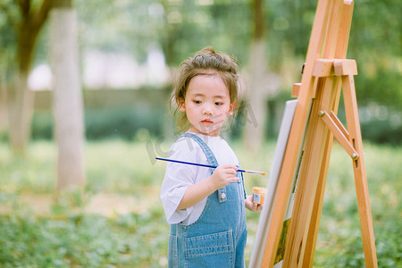 艺术启蒙白天穿着背带裤的小女孩户外草坪画画摄影图配图