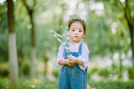 拿笔的卡通小女孩摄影照片_儿童玩耍白天可爱小女孩小树林吹泡泡摄影图配图