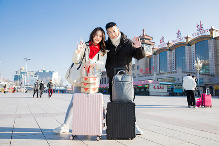 的行李箱摄影照片_春运白天拉着行李箱的年轻情侣火车站招手摄影图配图