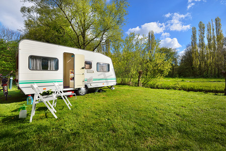 露营地绿色草坪上的白色大篷车拖车。阳光灿烂的日子。春天的风景。欧洲。生活方式、旅游、生态旅游、公路旅行、旅行、度假、娱乐、交通、旅行车、汽车回家