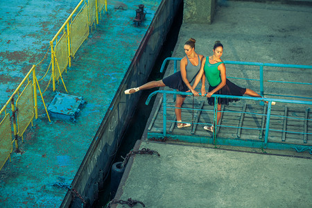 欧美服装背景摄影照片_两个年轻漂亮的双胞胎姐姐在城市与芭蕾舞服装跳芭蕾舞。城市同步舞蹈。工业街舞。具有特殊效果。同步构成。普安特芭蕾舞鞋和衣服.