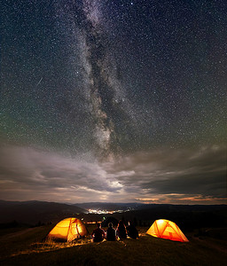 四名徒步旅行者坐在两个橙色帐篷营地, 晚上享受美丽的星空和银河。在远处的城市的灯光坐落在山之间