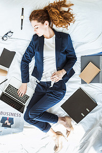 女躺摄影照片_在文件夹和文档中躺在床上时, 在笔记本电脑上打字的女商人的最高视图