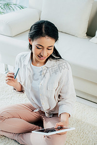 微笑的拉丁女人拿着信用卡，使用数字平板电脑，而坐在地板上在家里