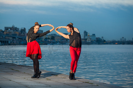 欧美服装背景摄影照片_在河附近城市的背景下，两个年轻漂亮的双胞胎姐妹在跳舞 waacking 舞蹈。在夏天时间上显示不同的风格和现代舞蹈与黑色和红色的衣服，近水的姿势.