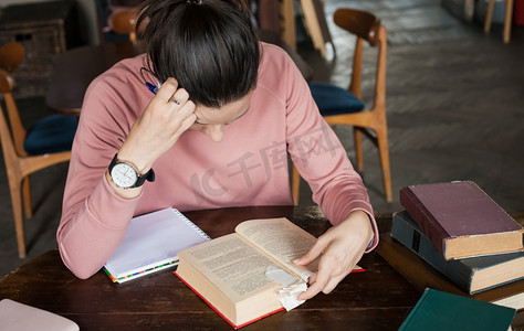 考试准备。戴着眼镜和粉红色毛衣的学生女孩靠在书上，在桌子上的一个旧图书馆里工作.