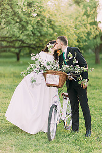 浪漫的年轻人刚刚结婚情侣接吻在阳光明媚的公园与装饰的自行车。温暖的太阳耀斑