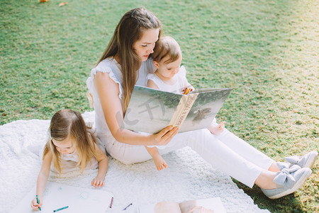 两个可爱的小妹妹和他们的母亲在公园的夏季野餐的图画和读书书