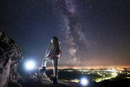 发光宇宙摄影照片_可以看到太空旅行者站在岩石山上，欣赏着星空、银河和夜城的美景。身穿白色宇航服的飞行任务专业宇航员。空间旅行的概念