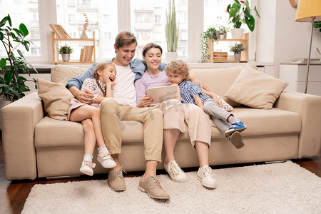 欧美生活摄影照片_快乐的孩子们和他们的父母都穿着便服，在客厅的沙发上放松，在平板电脑上看卡通片