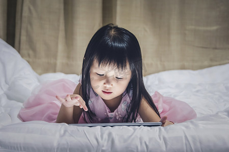 小女孩躺在床上晚上使用互联网观看电视或 gamp 玩儿童智能手机或片剂上瘾概念.