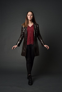 穿红色长皮革插孔的黑发女孩的全长肖像。站在灰色工作室背景的姿势.