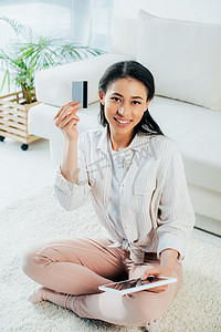 credit摄影照片_attractive young woman using digital tablet and holding credit card while sitting on floor and smiling at camera