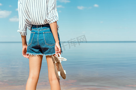 在海前手持凉鞋的年轻女子的裁剪图像 