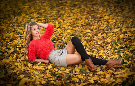 与红色上衣和短裙，在秋天期间在公园里摆美丽优雅的女人。年轻漂亮的女人，一头金发，躺在秋天的叶子。性感的金发女郎，有着黑色的紧身裤，在森林里.