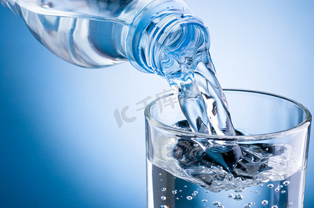 蓝色饮料摄影照片_倒水从瓶子变成蓝色背景上的玻璃