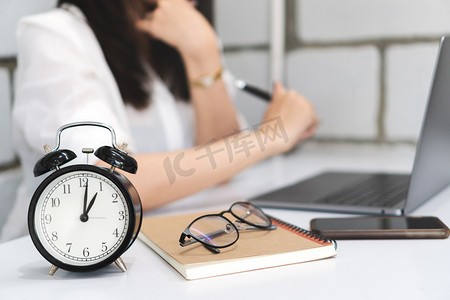 时间管理, 关闭黑色闹钟与模糊的背景的商业妇女正在使用笔记本电脑, 智能手机, 书籍, 钢笔和眼镜在办公桌上的办公室。工作时间概念.