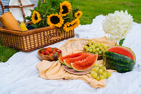 夏日野餐的概念,阳光灿烂,有西瓜、水果、花束、水仙花和向日葵花.带着食物的野餐篮和白色针织毛毯的令人神清气爽的夏季饮料。有选择的重点