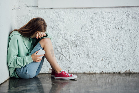 悲伤摄影照片_悲伤的哭泣的女孩坐在纹理墙附近，拿着智能手机