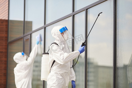 两名穿着防护服的工人在建筑物室外消毒或清洁、复制空间时喷洒化学品的肖像
