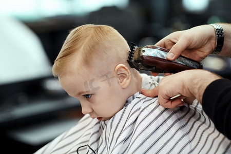 可爱的金发男婴与蓝眼睛在理发店理发由理发师。造型师的手与工具。儿童时尚在沙龙。室内，深色背景，复制空间.