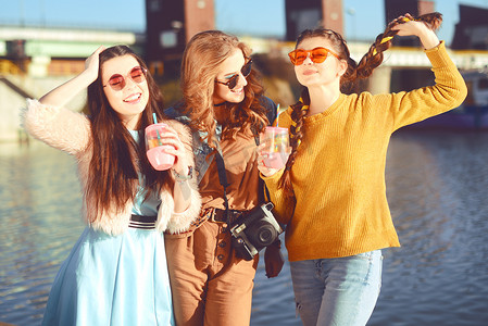 三个女孩在河边对着天空玩得很开心。戴着太阳镜的时尚女孩跳, 高兴, 喝酒, 微笑有乐趣, 疯了。穿着色彩的朋友花夏天的气氛, 时间在一起.
