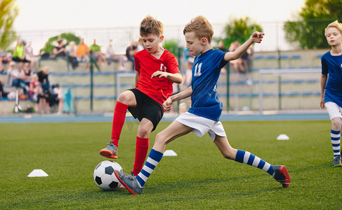 儿童足球运动员在足球场上踢球。体育足球横向背景。观众在后台体育场观看。身穿红衣和蓝衣的青少年运动员。体育教育