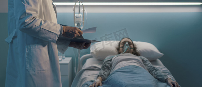 一名专业医生帮助躺在医院病床上的病人，他戴着氧气面罩，正在检查病历