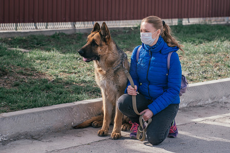 一个戴着防护医疗面具的女孩在街上遛狗。检疫期间和宠物一起休息.和一个德国牧羊人在新鲜空气中散步.自我隔离和保护模式.