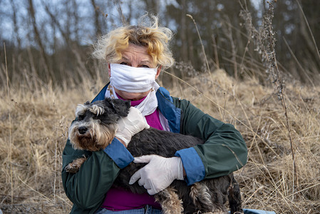 一位戴着防护医疗面罩和橡胶手套带着狗散步的老年妇女，一个小型施瑙策人.