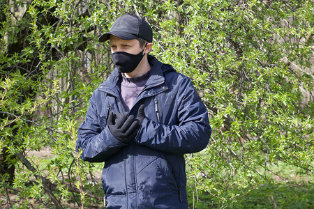 一个年轻人走过一个春天的公园，摆出姿势。他脸上戴着一个黑色的布面具.穿着一件春季夹克，头戴帽子。大流行病期间在新鲜空气中散步.