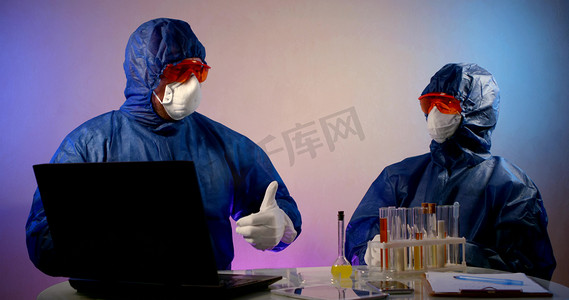 Two virologists are in the lab, sitting at a table with a laptop and flasks of analysis. They are wearing a blue work suit, glasses, and a mask. They are happy to work successfully