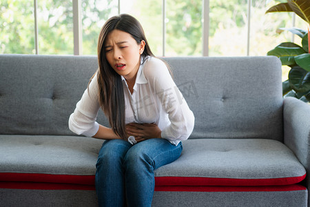 不快乐的亚洲女人坐在沙发上，忍受着肚子痛。月经、腹泻或消化不良引起的腹痛。疾病和保健概念