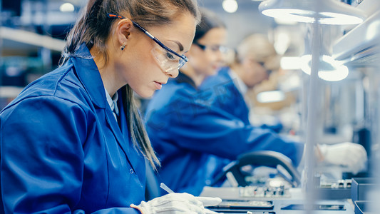 身穿蓝色工作服和保护眼镜的女性电子工厂工人用Tweezer组装智能手机的印刷电路板。拥有更多员工背景的高科技工厂.