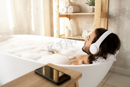 年轻男子头戴耳机，躺在布满热水和泡沫的白色浴缸里，享受着悠闲的音乐