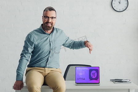 handsome businessman sitting on table and pointing with finger at laptop with web-shopping website 